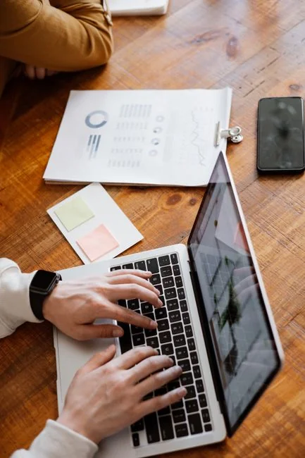 Des mains tapent sur un ordinateur portable tout en tenant un smartphone, illustrant un environnement de travail multitâche.
